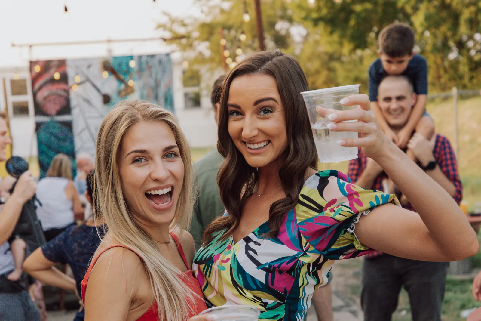 Zwei Freundinnen lachen und heben ihre Becher auf einer Gartenparty unter Lichterketten